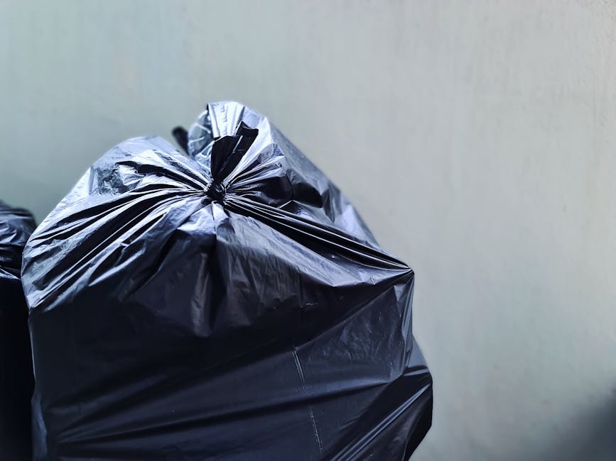 Close-up image of a tightly tied black plastic rubbish bag with a shiny, smooth surface, positioned on the left side of the frame. The bag appears full and is resting against a plain, light-colored wa