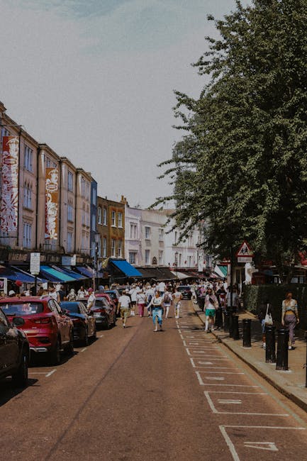 A busy street scene in an urban area with multi-storey buildings on the left, featuring facades in pastel shades of pink, yellow, and white, some with visible signage for shops and cafes. The street i